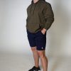 Forge Olive 4 A man stands against a plain light background, wearing an olive green hoodie, navy blue shorts, and black sneakers. His hands are in his pockets, and he looks slightly upward with a neutral expression.