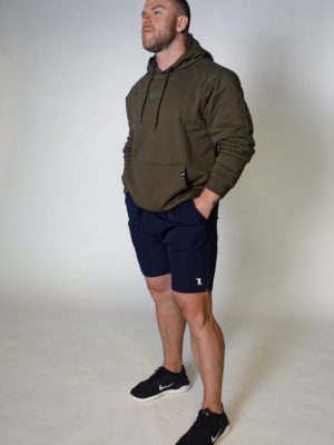 A man stands against a plain light background, wearing an olive green hoodie, navy blue shorts, and black sneakers. His hands are in his pockets, and he looks slightly upward with a neutral expression.
