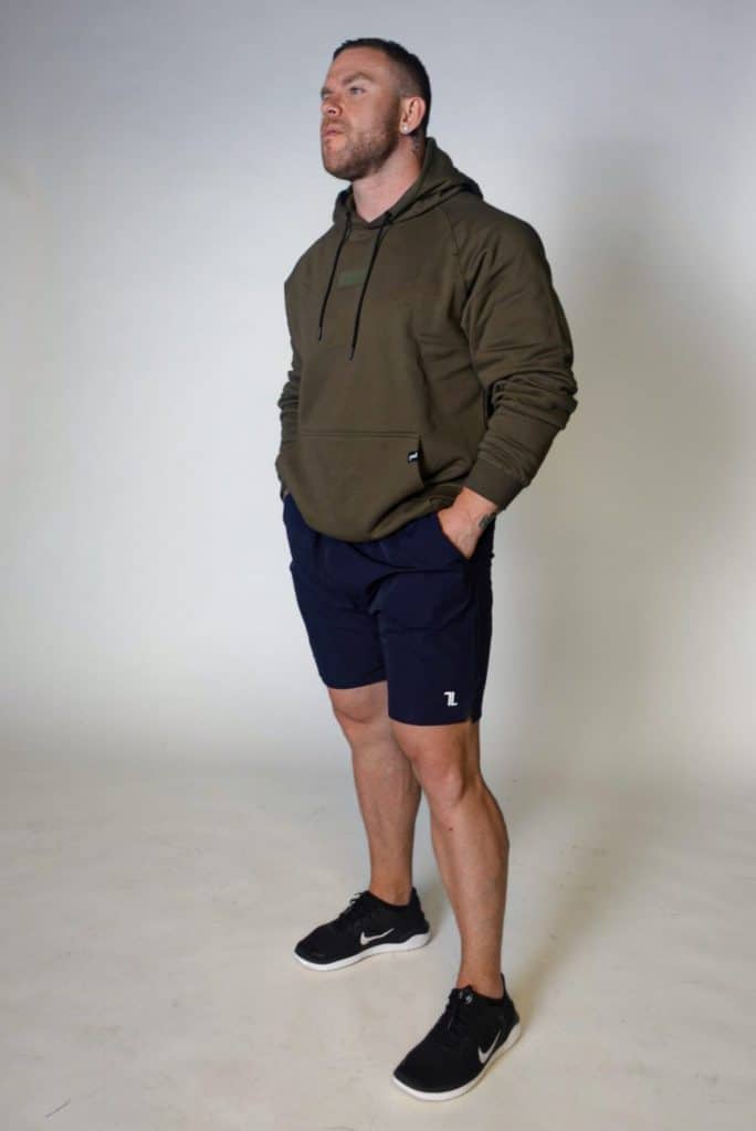 A man stands against a plain light background, wearing an olive green hoodie, navy blue shorts, and black sneakers. His hands are in his pockets, and he looks slightly upward with a neutral expression.
