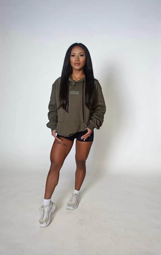A woman stands against a plain white background, wearing a green hoodie, black shorts, white socks, and white sneakers. She has long straight hair and a tattoo on her right thigh.