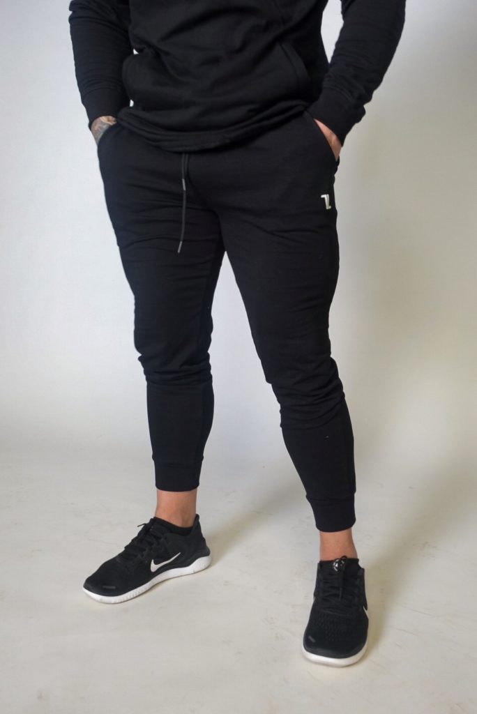 A person stands against a plain background, hands in pockets, wearing black Genesis Joggers, a black hoodie, and black Nike sneakers with white soles. Only the body from shoulders down is visible.