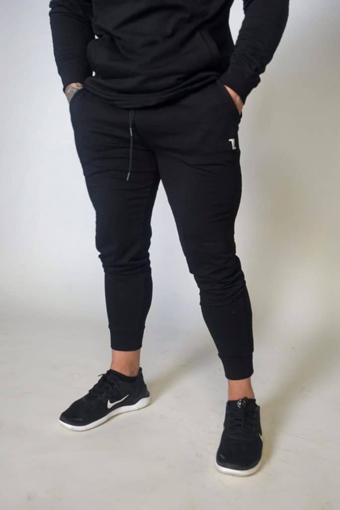 A person stands against a plain background, hands in pockets, wearing black Genesis Joggers, a black hoodie, and black Nike sneakers with white soles. Only the body from shoulders down is visible.