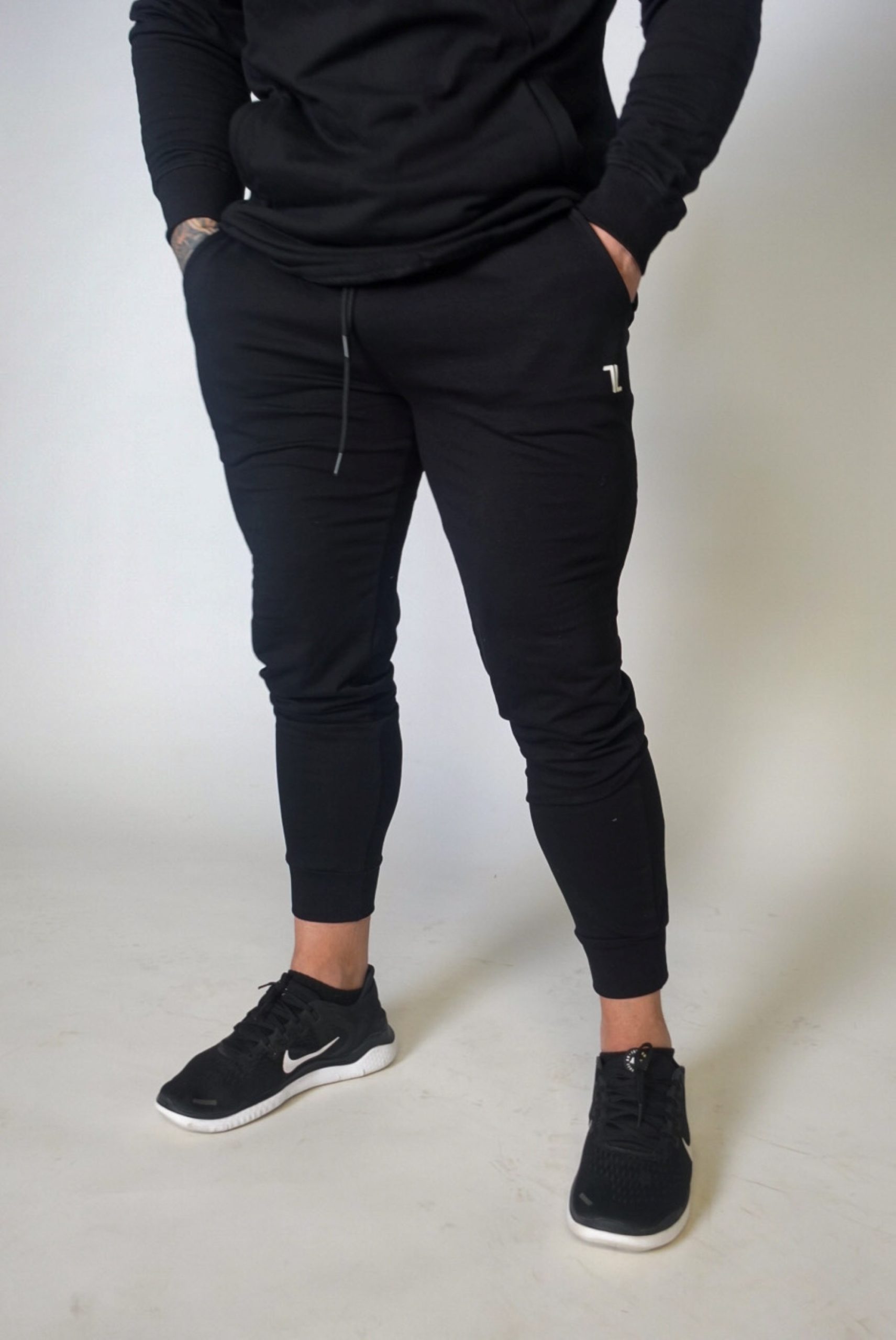 Wearing Genesis Joggers and a black hoodie, the person stands with hands in pockets against a plain light background. Black Nike sneakers complete the look; their face remains unseen.