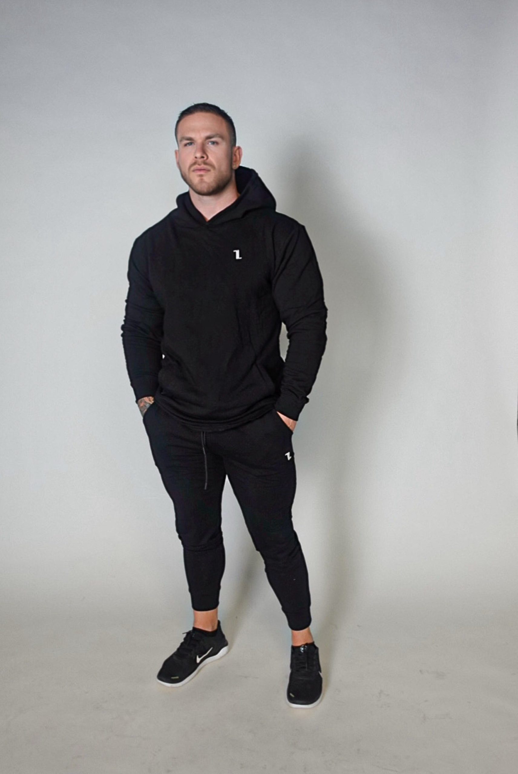 A man wearing black Genesis Joggers, a black hoodie, and black sneakers stands with his hands in his pockets against a plain light grey background.
