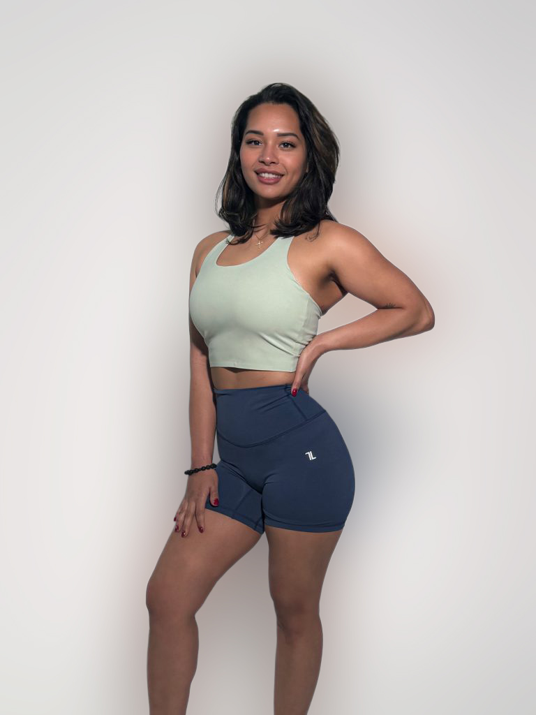 A woman smiles against a plain background wearing the FlowEase BraTop in light green and navy blue high-waisted athletic shorts, with one hand on her hip and the other on her thigh.
