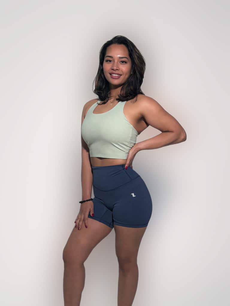 A woman smiles against a plain background wearing the FlowEase BraTop in light green and navy blue high-waisted athletic shorts, with one hand on her hip and the other on her thigh.