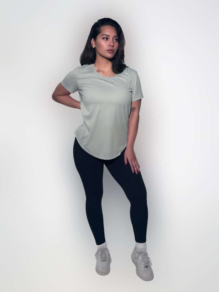 A woman stands against a white background, wearing the VeloraFlow Tee in light gray, paired with black leggings and white sneakers. She has one hand on her hip and looks confidently ahead.