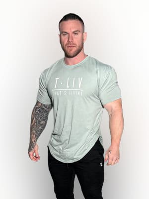 A muscular man with a beard stands against a white background, wearing a light gray "T.LIV THAT'S LIVING" t-shirt and black pants. He has a full sleeve tattoo on his left arm and a serious expression.