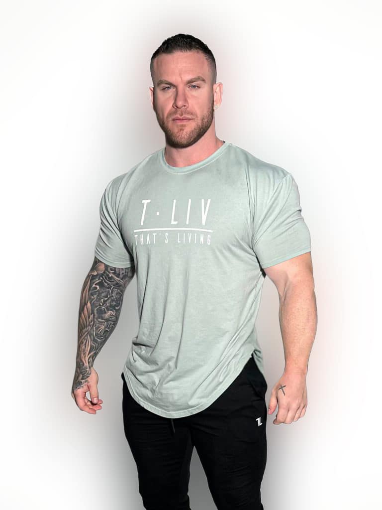 A muscular man with a beard stands against a white background, wearing a light gray "T.LIV THAT'S LIVING" t-shirt and black pants. He has a full sleeve tattoo on his left arm and a serious expression.