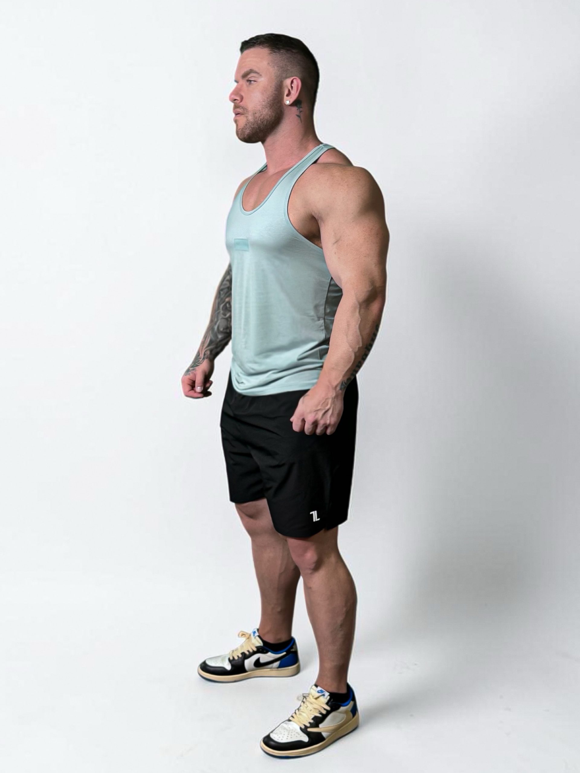 A muscular man with short hair stands in profile against a white background, wearing a light blue tank top, Rush Shorts, and sneakers. Tattoos are visible on his left arm.