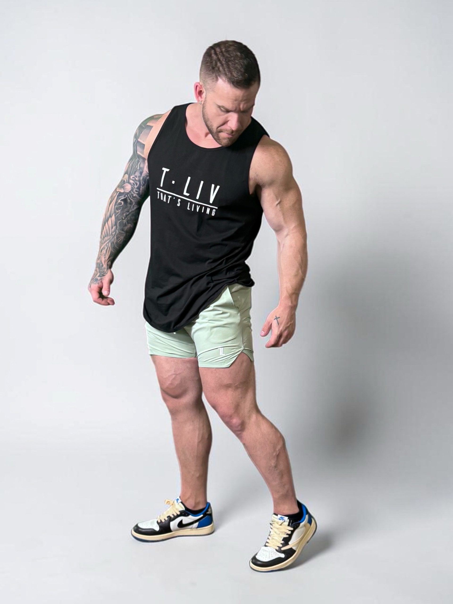 A muscular man with tattoos wears a black tank top that says "T.LIV THAT'S LIVING," light green shorts, and sneakers. He stands against a plain gray background, looking down to his left.