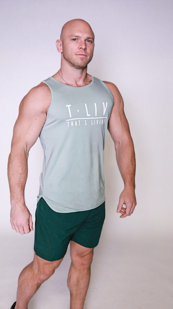A muscular bald man stands confidently against a plain light background, wearing the Genesis Tank—a light green sleeveless shirt with "T.LIV THAT'S LIVING" text—and dark green shorts.