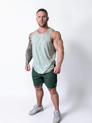 A muscular man with short hair stands confidently against a plain white background, wearing a light green sleeveless tank top, dark green shorts, and gray athletic shoes. One arm features a large tattoo.