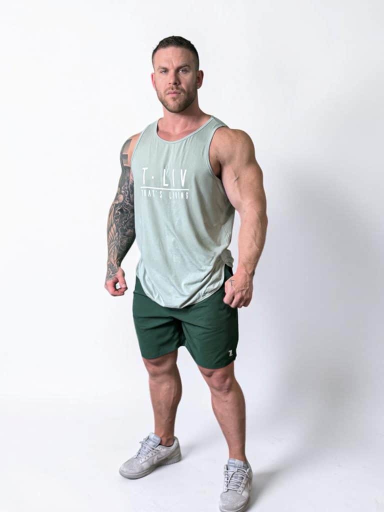 A muscular man with short hair stands confidently against a plain white background, wearing a light green sleeveless tank top, dark green shorts, and gray athletic shoes. One arm features a large tattoo.