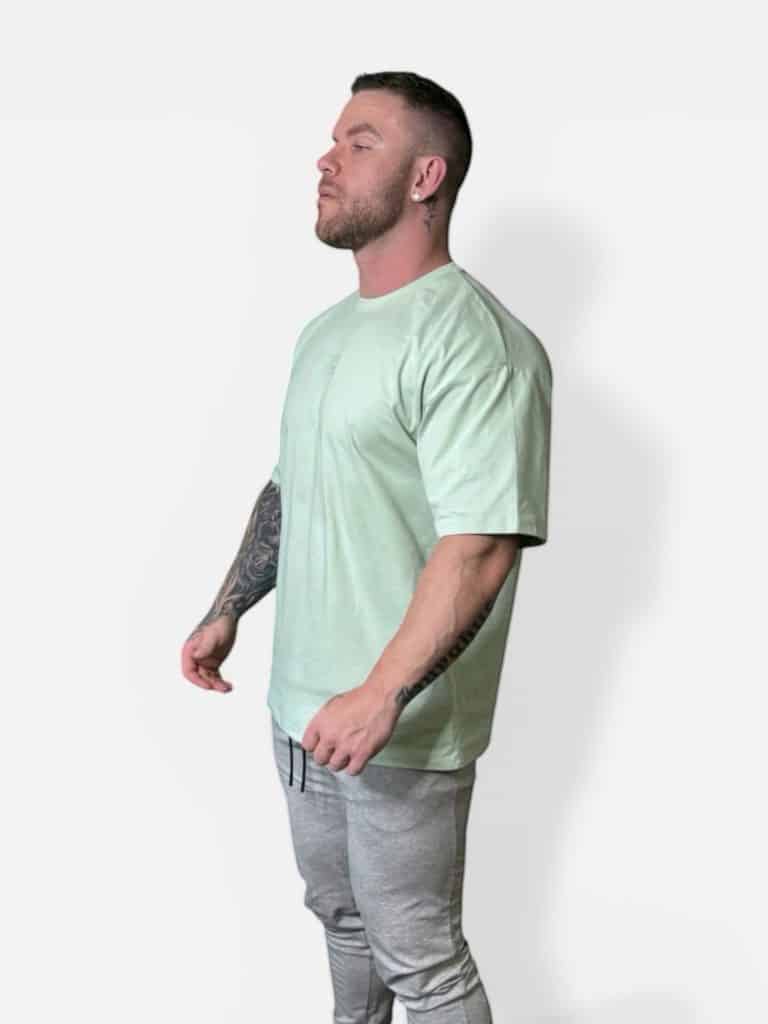 A man with tattoos on his left arm is standing in profile against a plain white background. He is wearing a light green T-shirt and light gray joggers.