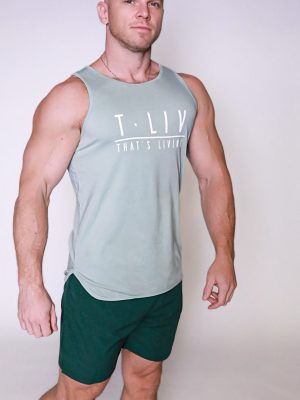 A muscular bald man stands confidently against a plain light background, wearing the Genesis Tank—a light green sleeveless shirt with "T.LIV THAT'S LIVING" text—and dark green shorts.