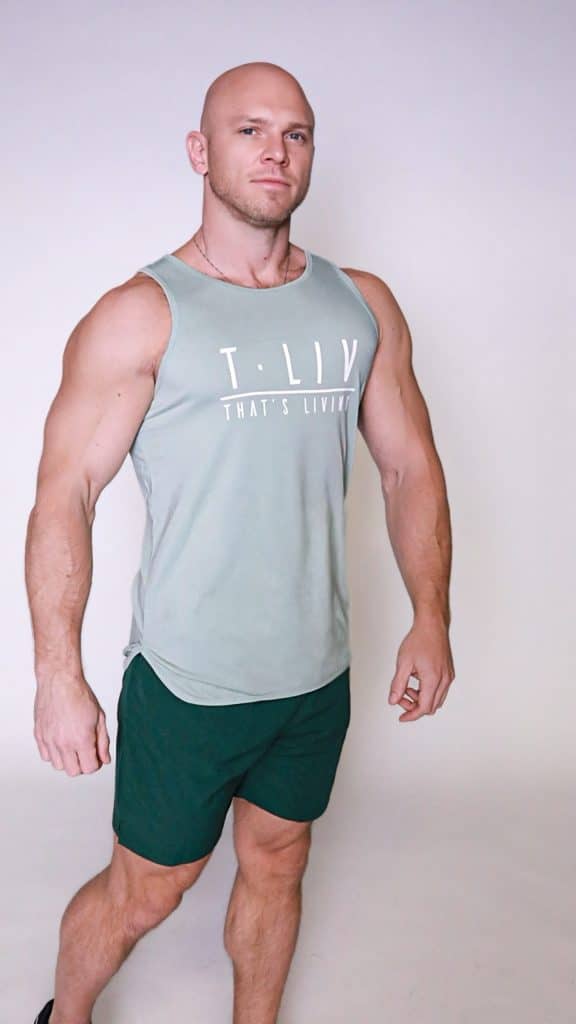 A muscular bald man stands confidently against a plain light background, wearing the Genesis Tank—a light green sleeveless shirt with "T.LIV THAT'S LIVING" text—and dark green shorts.