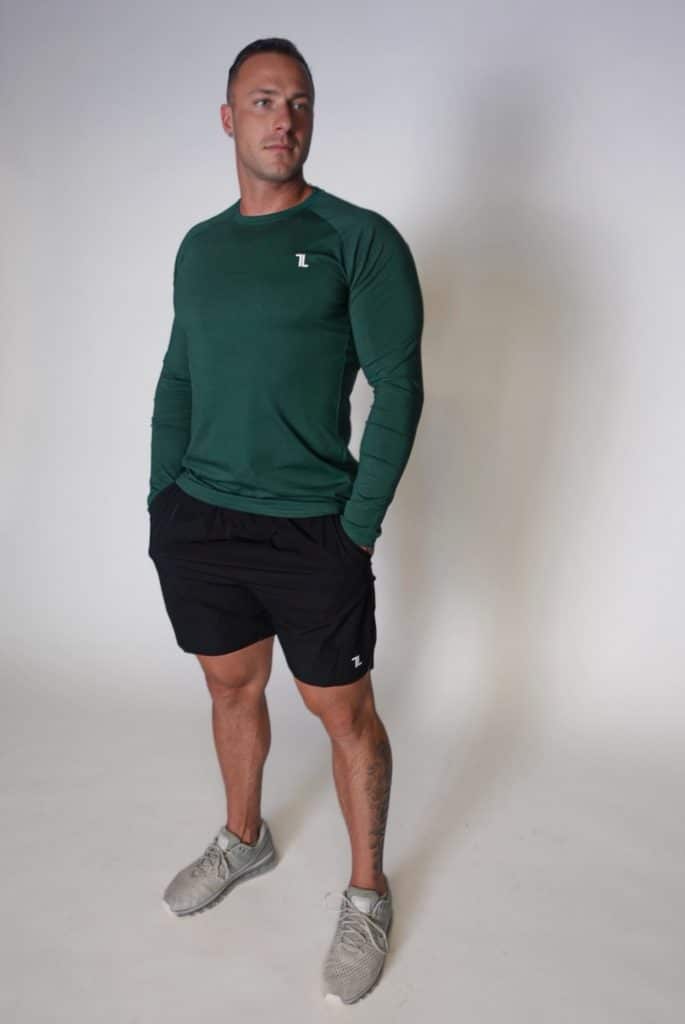 A man in a green long-sleeve athletic shirt, black shorts, and gray sneakers stands against a plain white background, looking to the side with his hands in his pockets.