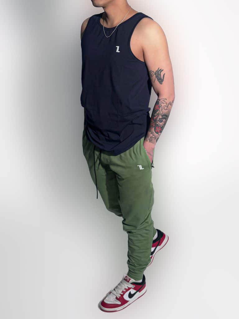 A person wearing a black sleeveless shirt, olive green joggers, and red-and-white Nike sneakers stands with hands in pockets. The person has a tattooed arm and is photographed against a plain white background.