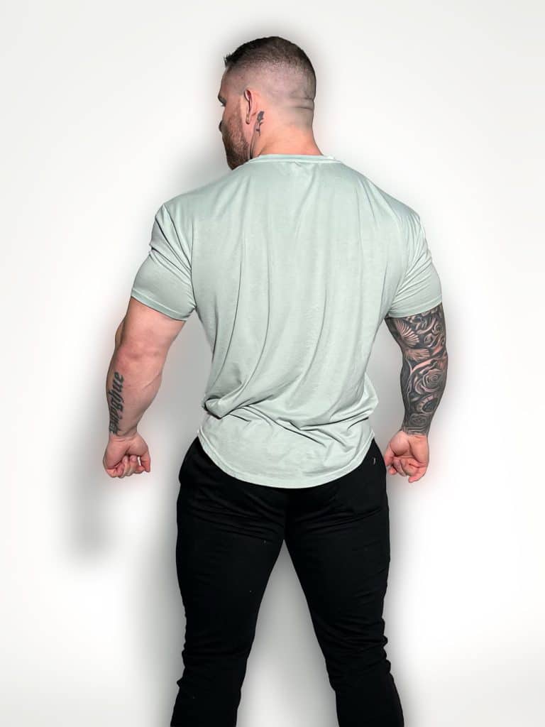 A muscular man with short hair stands facing away from the camera, wearing a light gray t-shirt and black pants. His left arm is heavily tattooed, and he is posing against a plain white background.