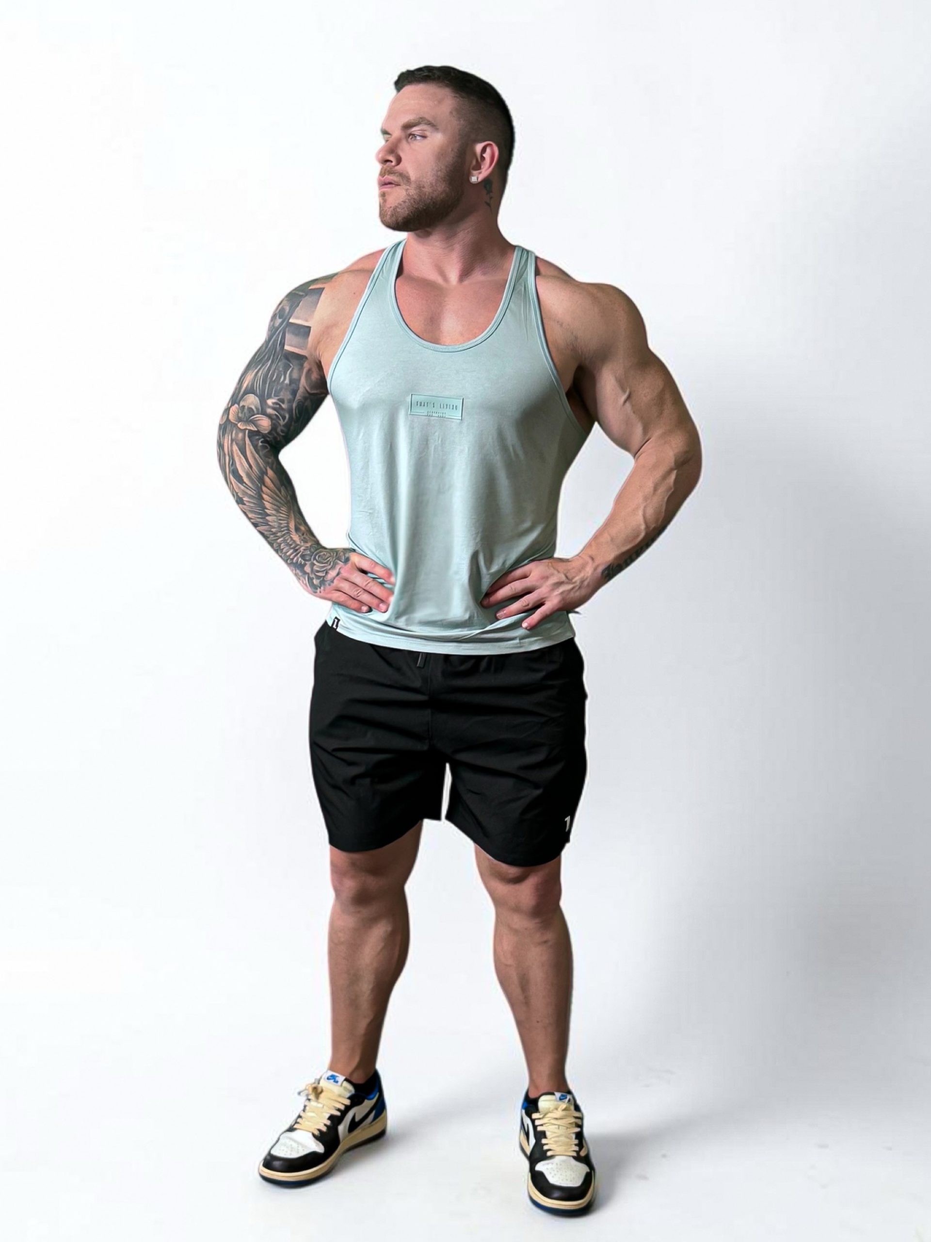 A muscular man with a tattoo sleeve on his left arm stands against a white background wearing Rush Shorts, a light blue tank top, and sneakers, hands on hips and looking to the side.