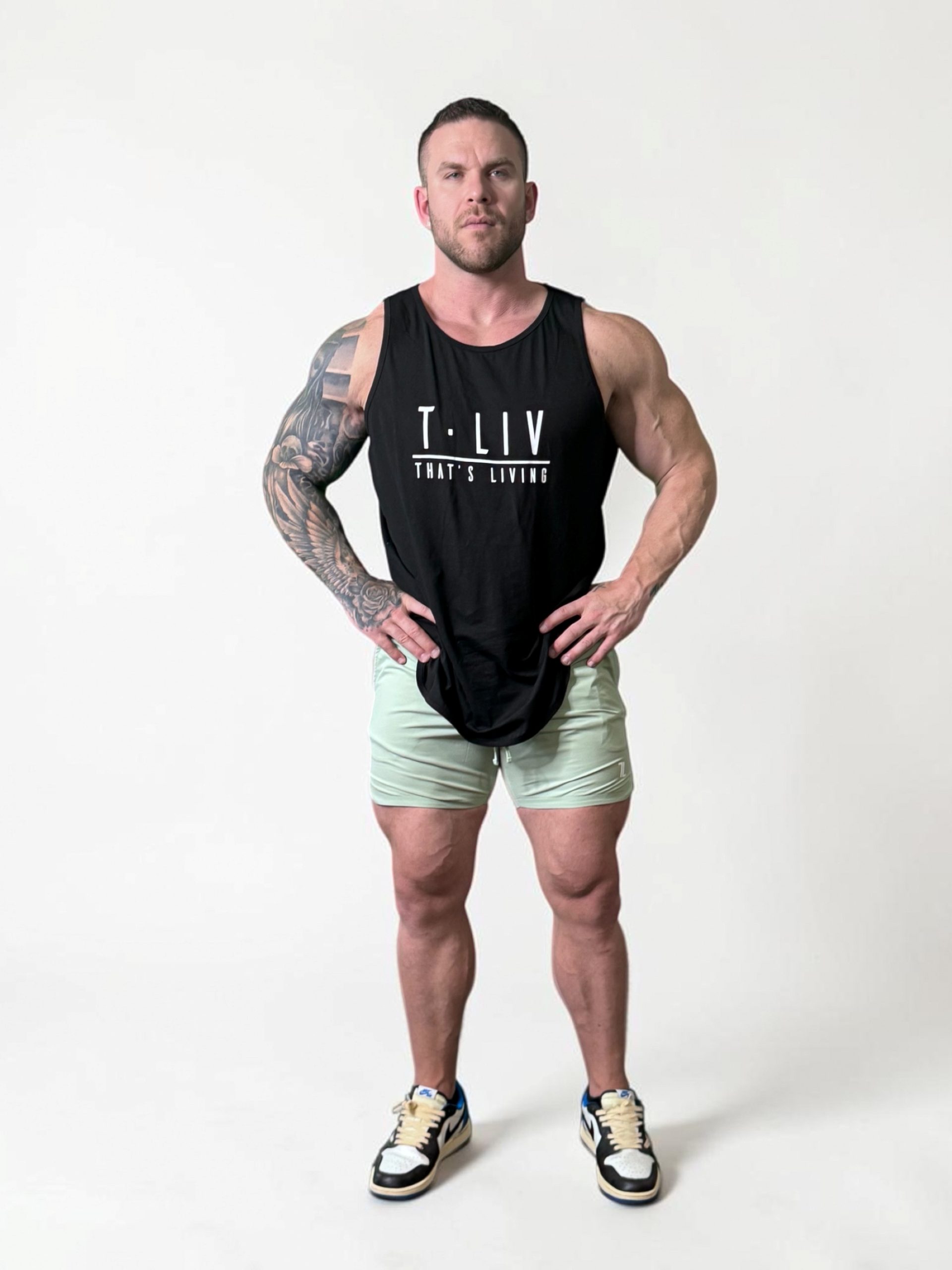 A muscular man with tattoos stands facing forward in a studio, wearing a black "T.LIV That's Living" tank top, light green shorts, and sneakers. He stands confidently with hands on hips against a plain white background.