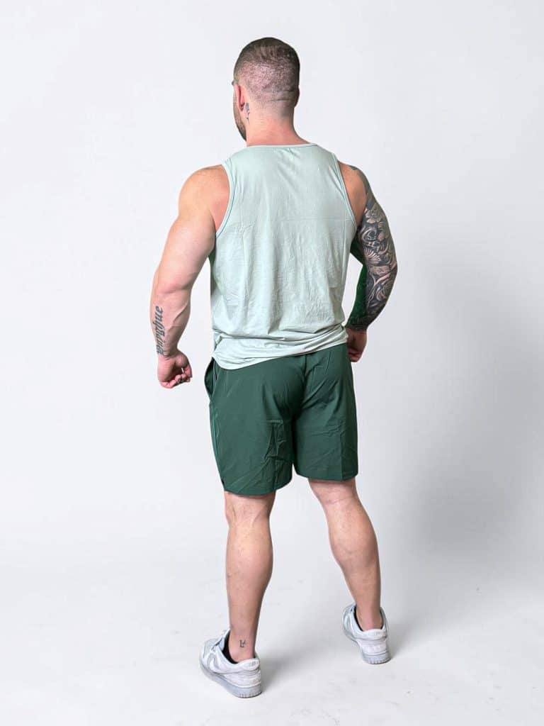 A muscular man stands facing away, wearing a light green sleeveless shirt, dark green shorts, and white sneakers. He has tattoos on his left arm and both calves, and stands against a plain white background.