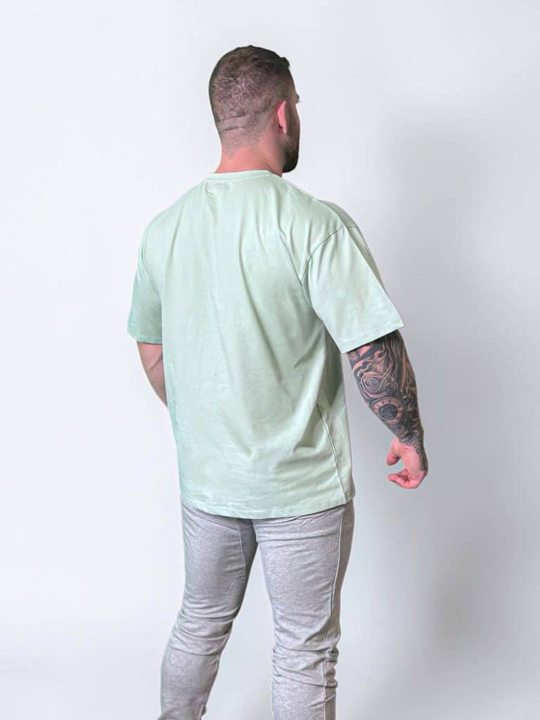 A man stands facing away from the camera, wearing a light green T-shirt and light gray pants. His right arm has a detailed tattoo sleeve, and he is standing against a plain white background.
