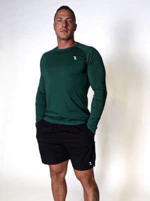 A man stands against a plain white background wearing a long-sleeve, dark green athletic shirt and black shorts. His hands are in his pockets, and he is facing forward with a neutral expression.