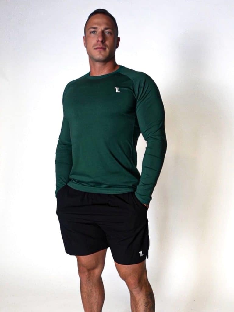A man stands against a plain white background wearing a long-sleeve, dark green athletic shirt and black shorts. His hands are in his pockets, and he is facing forward with a neutral expression.