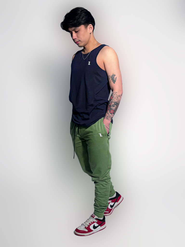 A man stands in a studio against a white background, wearing a navy tank top, green joggers, red and white Nike sneakers, and a necklace. He looks down with his hands in his pockets, showing a tattooed arm.