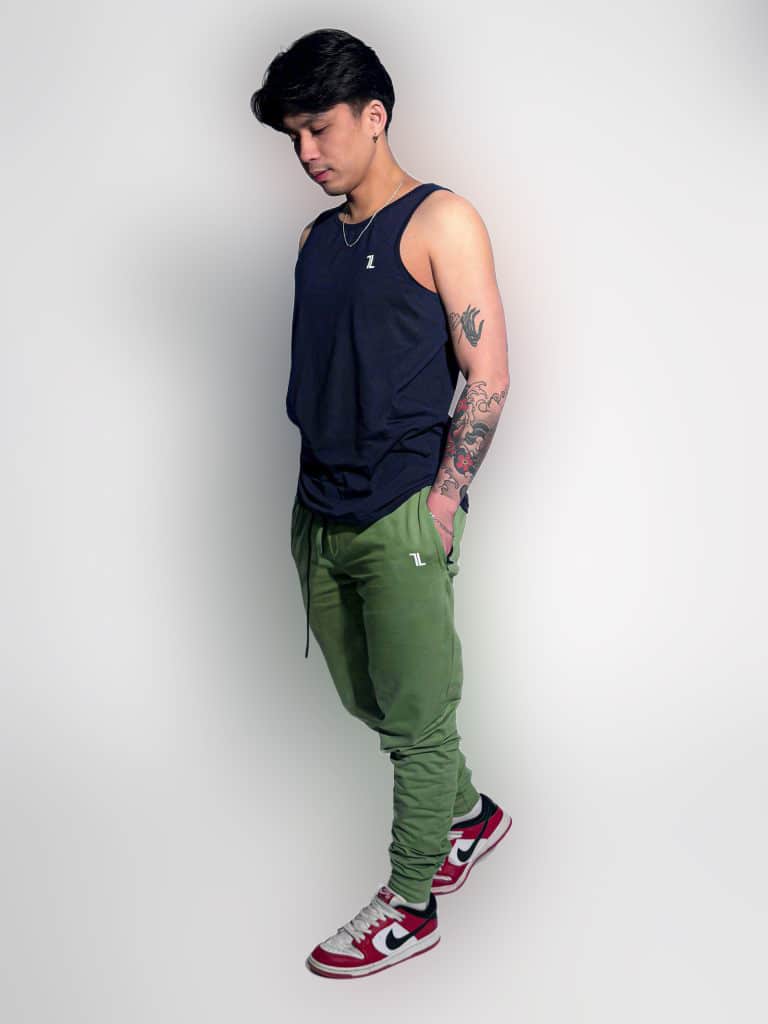 A man stands in a studio against a white background, wearing a navy tank top, green joggers, red and white Nike sneakers, and a necklace. He looks down with his hands in his pockets, showing a tattooed arm.