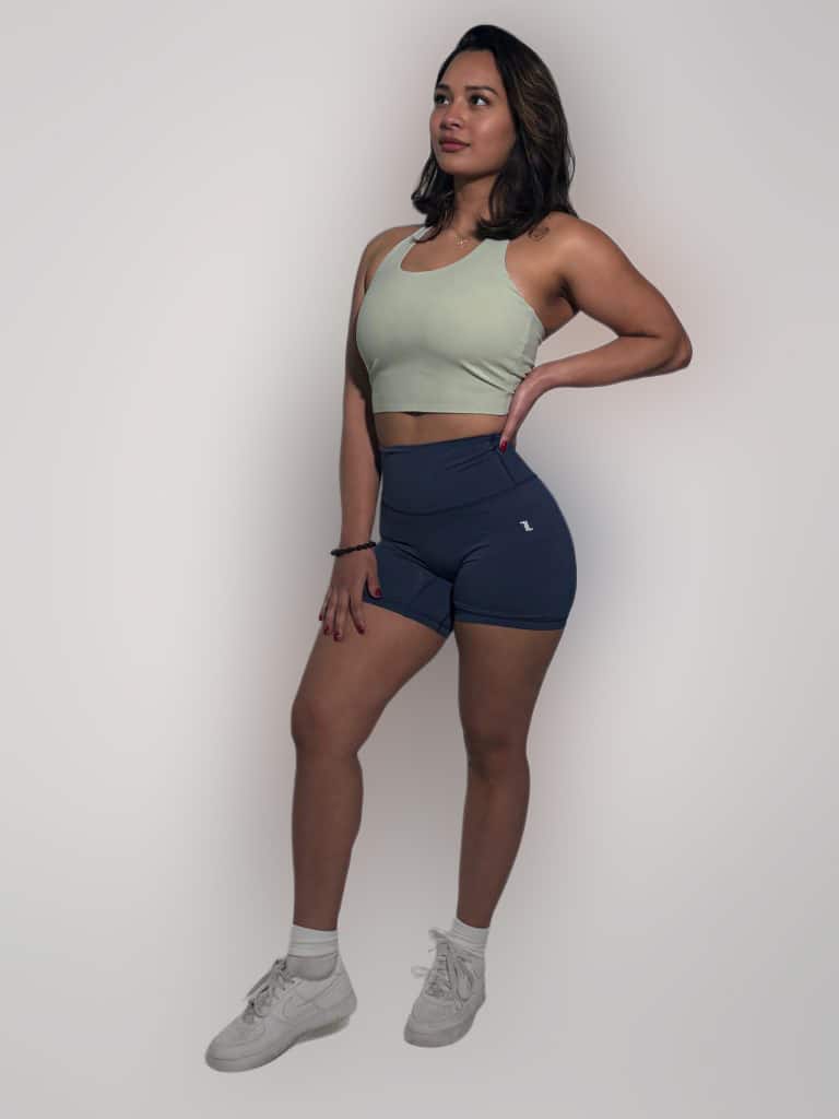 A woman with shoulder-length hair stands confidently against a plain background, wearing the FlowEase BraTop in light green, blue athletic shorts, white socks, and white sneakers. Her hand rests on her hip.