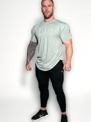 A muscular man with a beard stands against a plain white background, wearing a light green T-shirt that says "T. LIV WHAT'S LIVING," black jogger pants, and black Nike sneakers. One arm features a detailed tattoo sleeve.