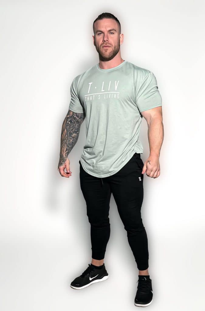 A muscular man with a beard stands against a plain white background, wearing a light green T-shirt that says "T. LIV WHAT'S LIVING," black jogger pants, and black Nike sneakers. One arm features a detailed tattoo sleeve.