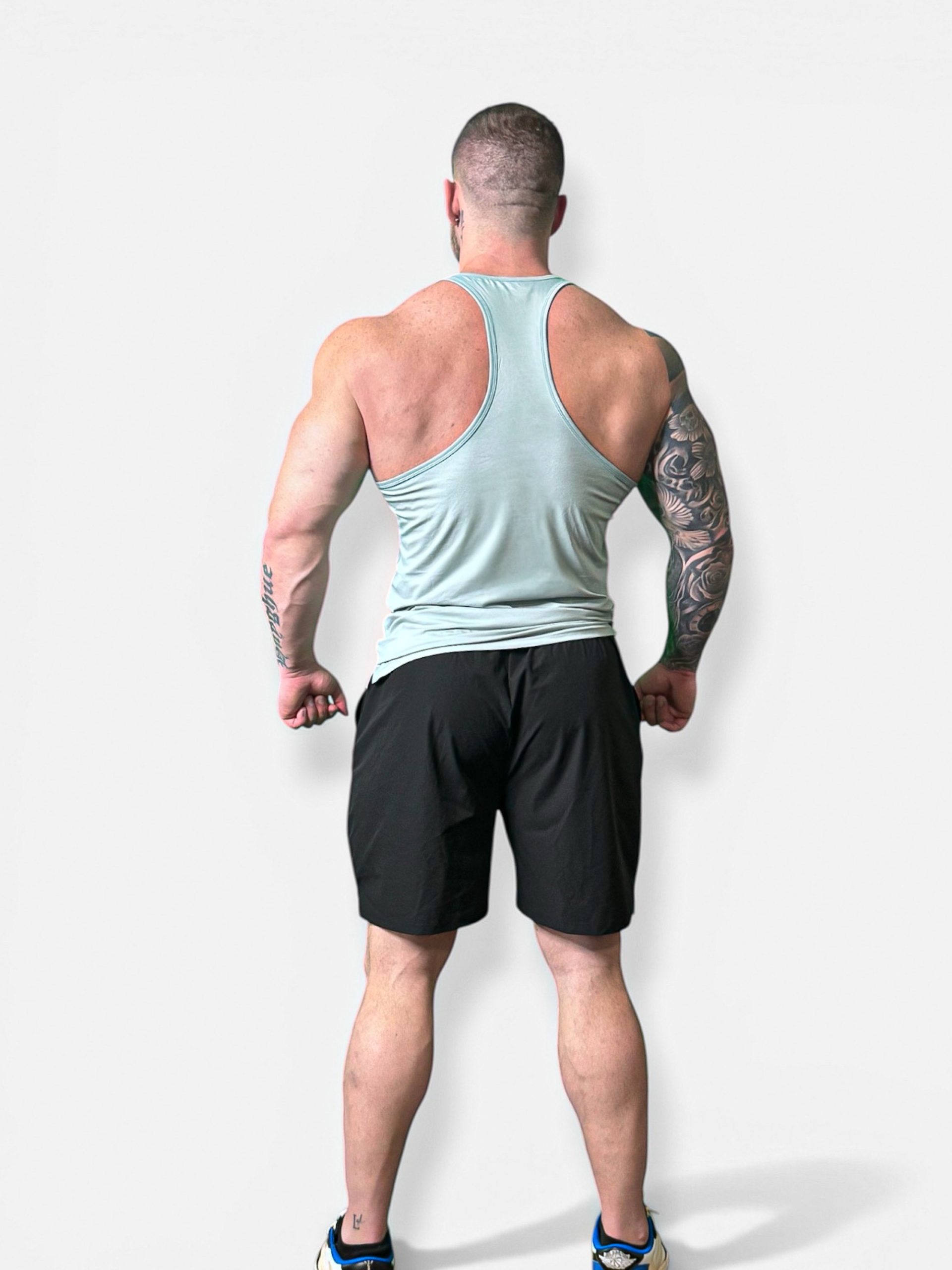 A muscular man faces away from the camera, wearing a light blue tank top, Rush Shorts, and sneakers. His arms are by his sides, displaying a tattoo sleeve on his right arm against a plain white background.