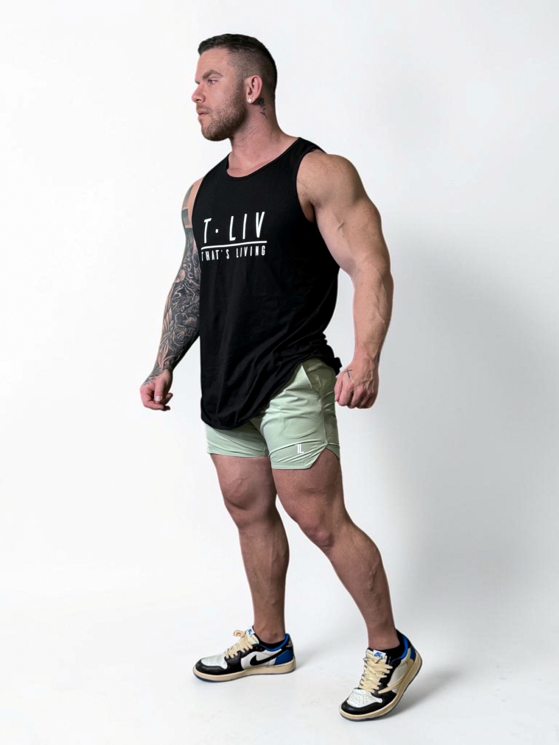 A muscular man with a tattooed right arm stands against a white background, wearing a black tank top, light green shorts, and white sneakers with black and blue details. He is looking to the left with a confident posture.