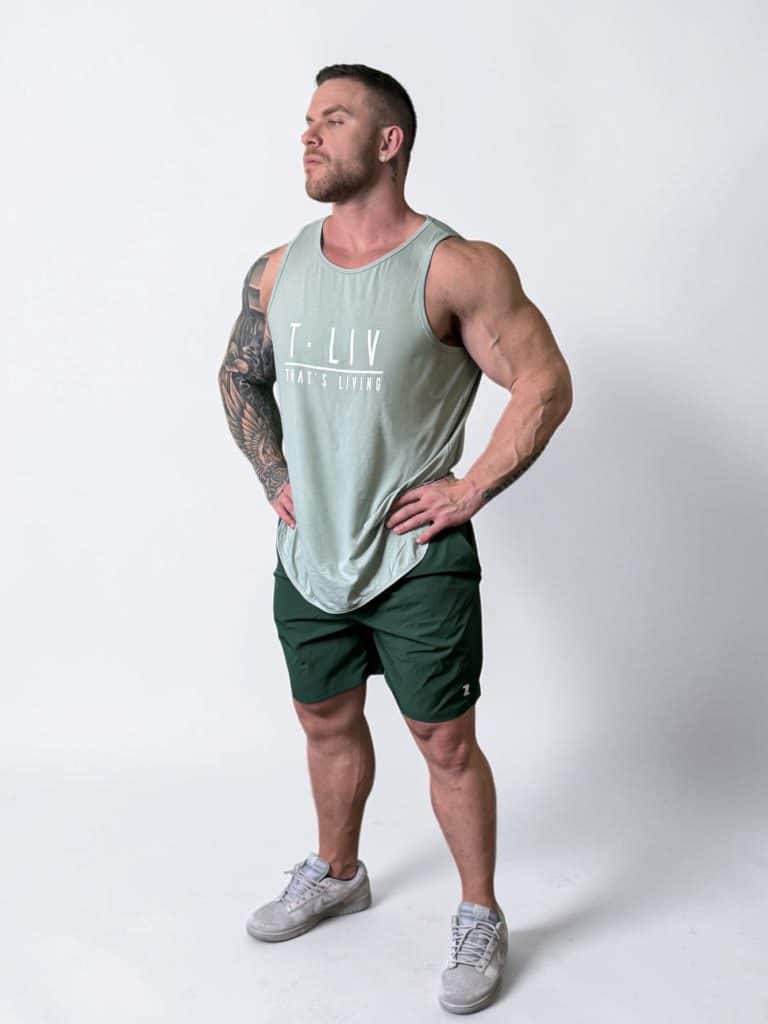 A muscular man wearing a light green sleeveless shirt, dark green shorts, and gray sneakers stands confidently with hands on hips against a plain white background.