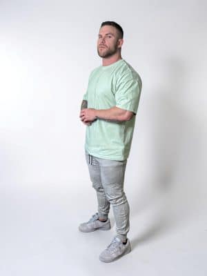 A man with short hair and a beard stands in a studio against a plain white background, wearing a light green T-shirt, gray jogger pants, and gray sneakers. He is looking slightly upward with his hands clasped.