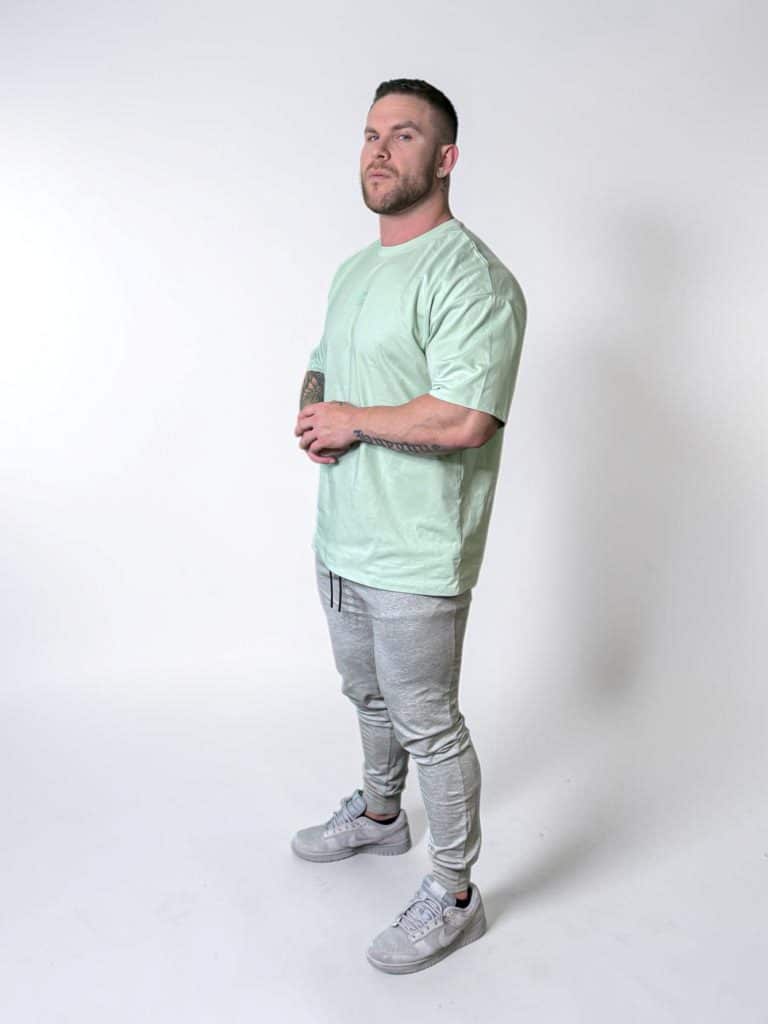 A man with short hair and a beard stands in a studio against a plain white background, wearing a light green T-shirt, gray jogger pants, and gray sneakers. He is looking slightly upward with his hands clasped.