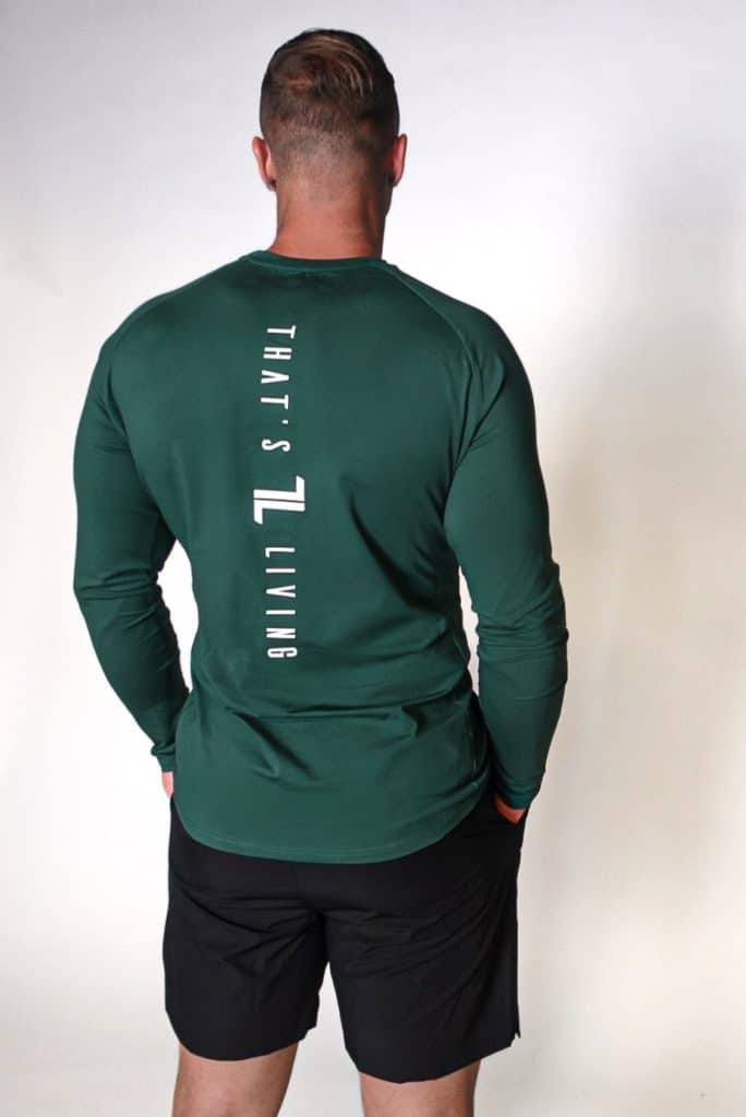 A man stands facing away from the camera, wearing a long-sleeve green shirt with "That's Living" printed vertically on the back, paired with black shorts, against a plain white background.