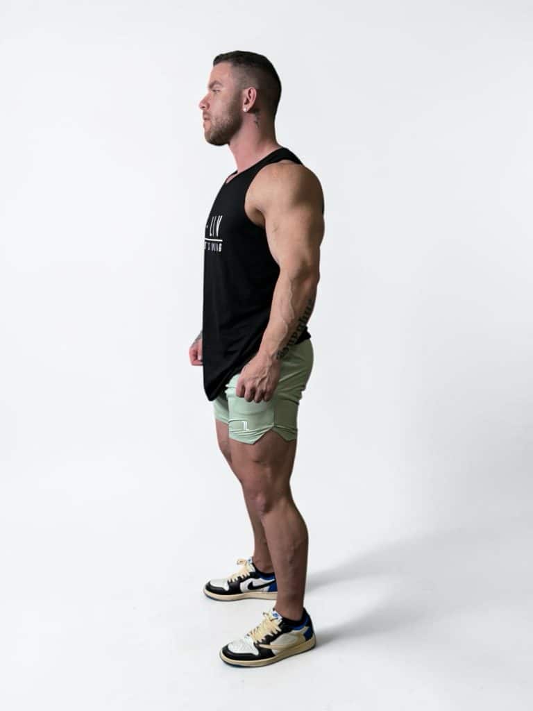 A muscular man stands in profile against a plain white background, wearing a black tank top, light green shorts, and black and white sneakers.