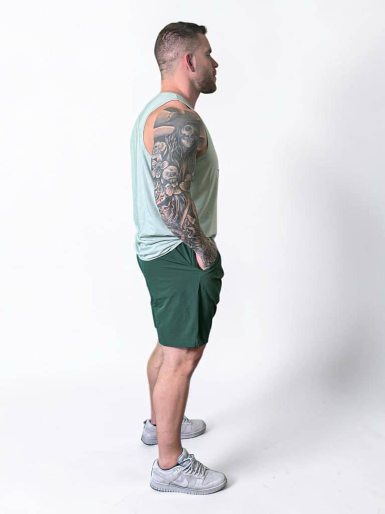 A man with a muscular build and a full sleeve tattoo on his left arm stands in profile, wearing a light green tank top, dark green shorts, and gray sneakers against a plain white background.