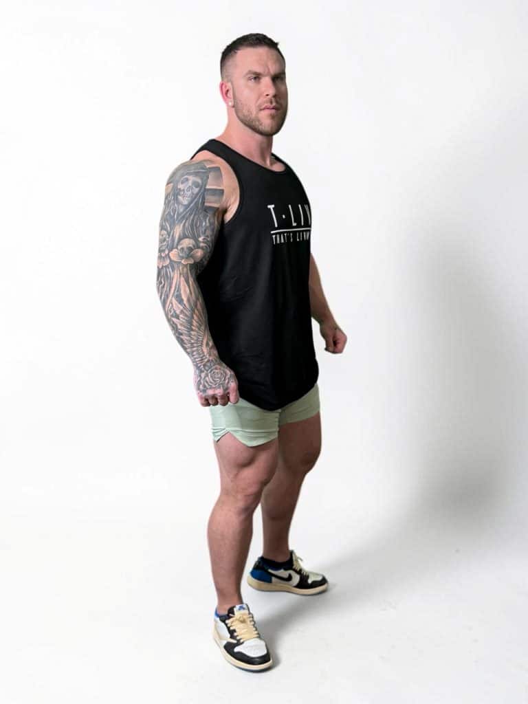 A muscular man with a large arm tattoo wears a black tank top, light green shorts, and sneakers, standing confidently against a plain white background.