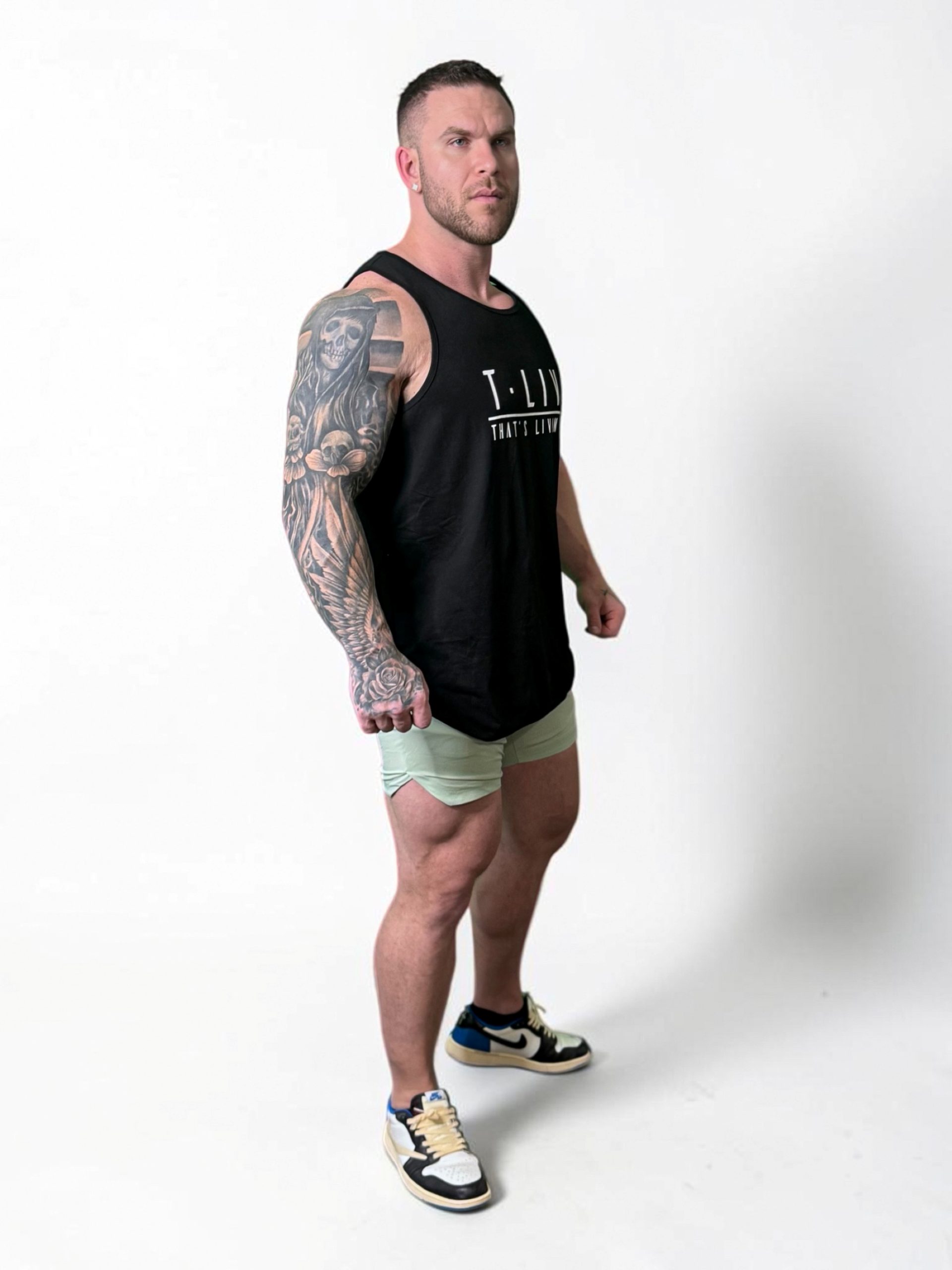 A muscular man with a large arm tattoo wears a black tank top, light green shorts, and sneakers, standing confidently against a plain white background.