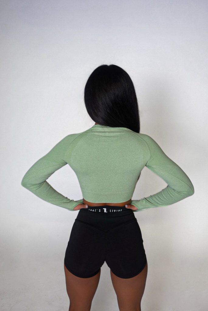 A woman with long dark hair, seen from behind, models the Allure Set V2—a light green long-sleeve crop top and black athletic shorts—against a plain white background.