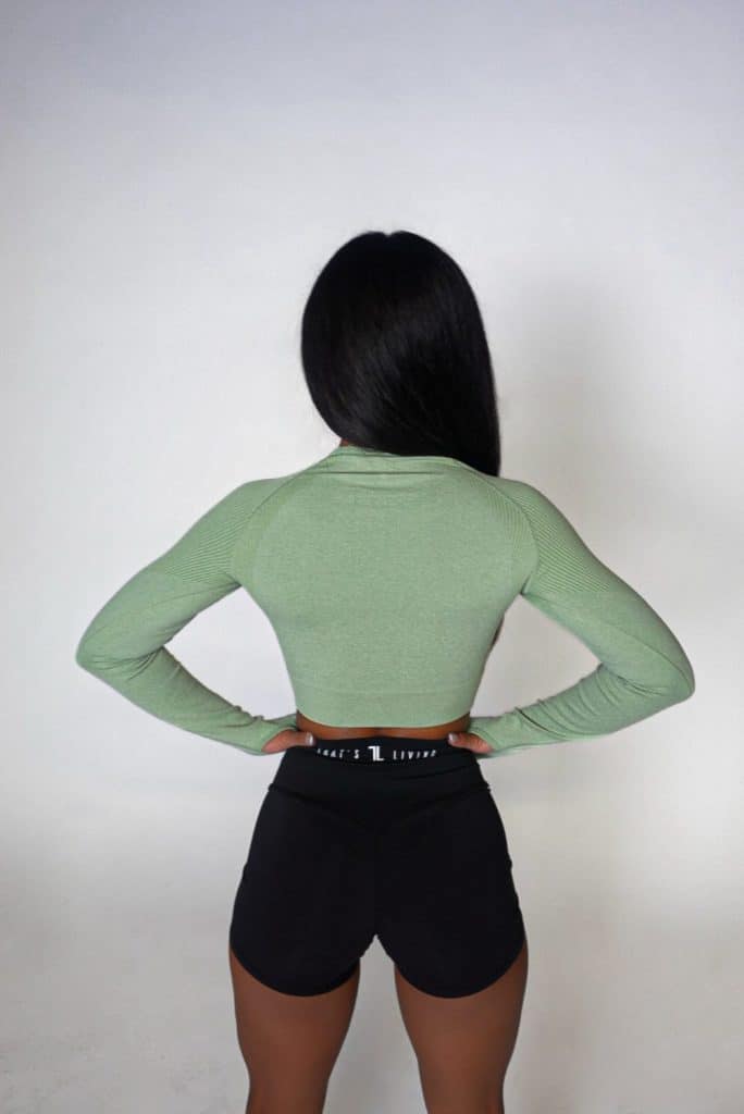 A woman with long dark hair, seen from behind, models the Allure Set V2—a light green long-sleeve crop top and black athletic shorts—against a plain white background.