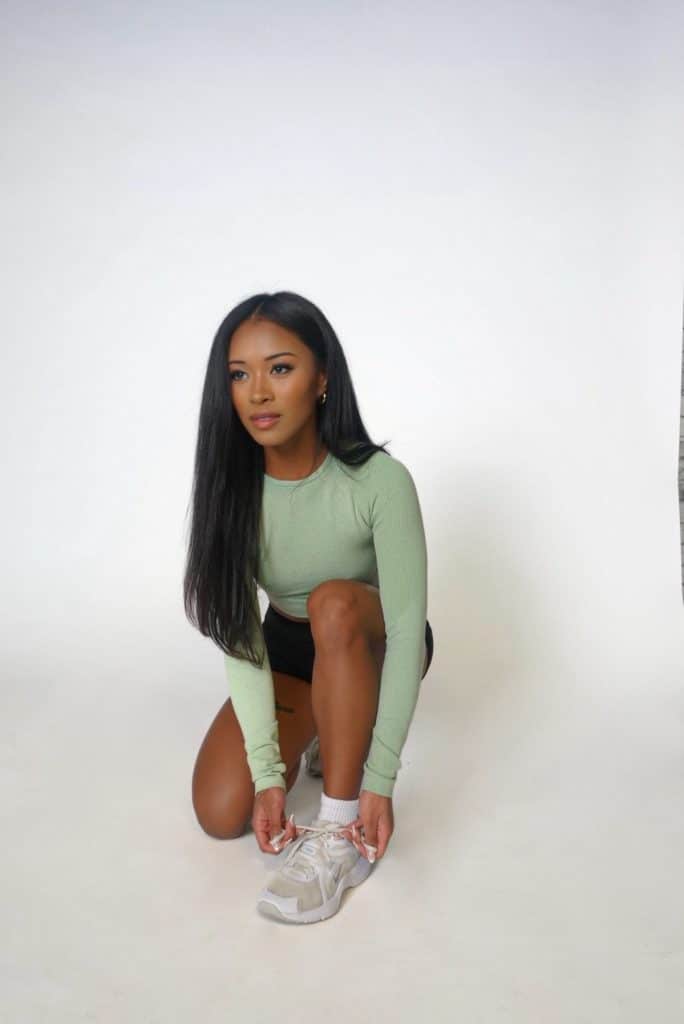 A woman with long, straight dark hair crouches to tie her white sneaker, dressed in the Allure Set V2—a light green long-sleeved top and black shorts—paired with white socks, against a plain white background.