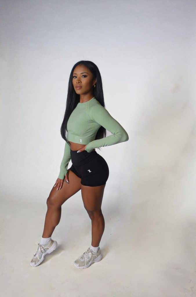 A woman with long straight hair confidently poses in the Allure Set V2—featuring a light green long-sleeve crop top, black shorts, and white sneakers—against a plain white background.
