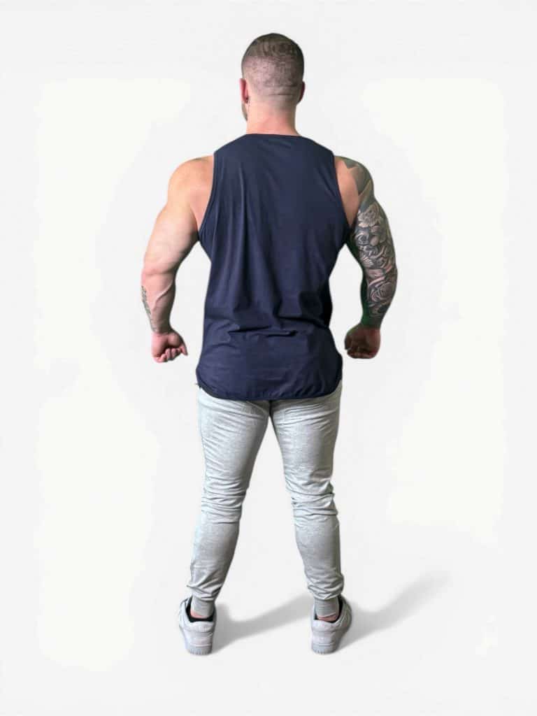 A muscular man with short hair, wearing Genesis Joggers in light gray, a sleeveless black shirt, and white sneakers, stands with his back to the camera, arms slightly bent and fists clenched.